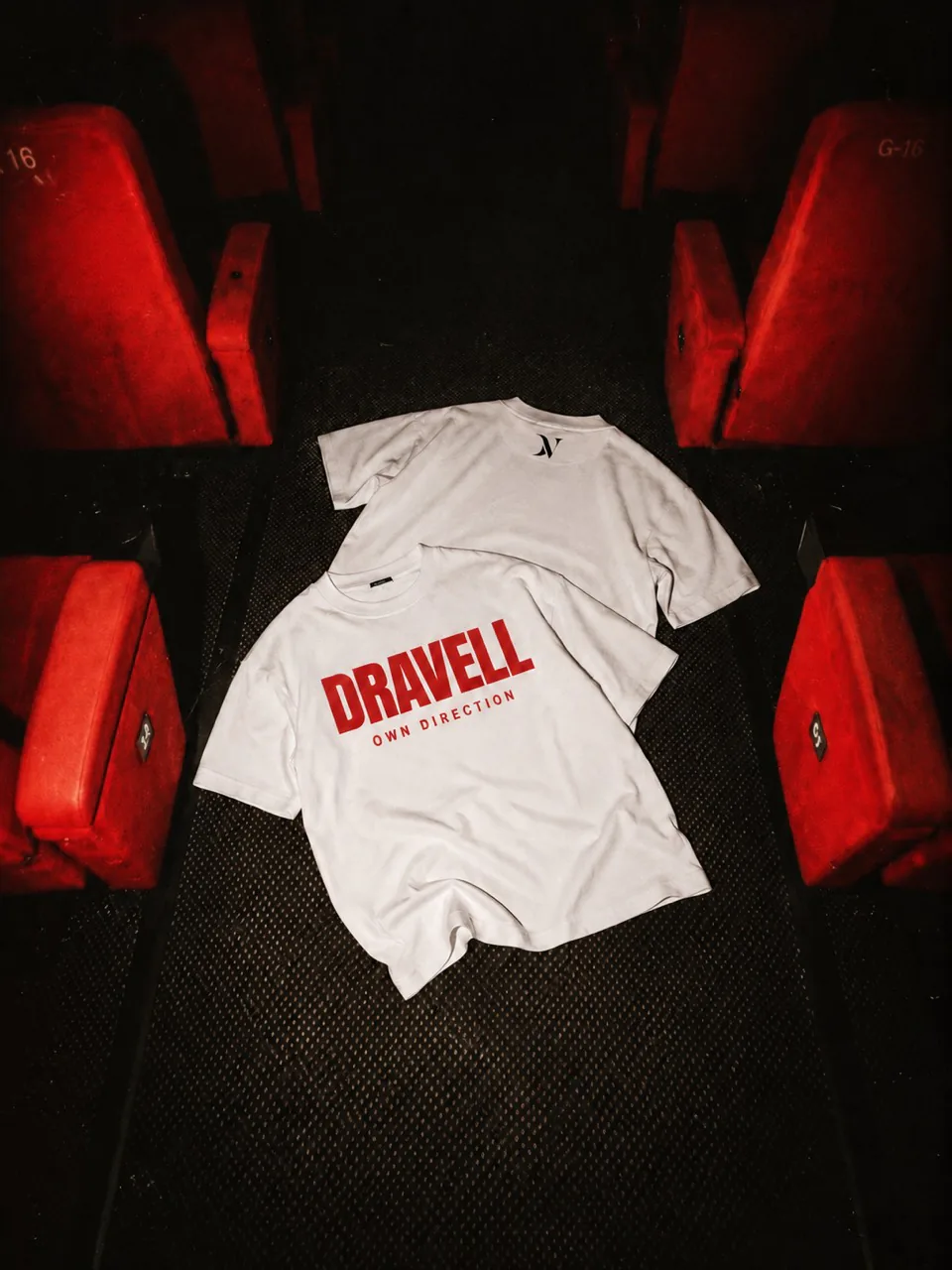 Des T-shirts DRAVELL blancs entre des sièges de cinéma rouges, avec lettrage rouge et petit logo noir au dos.