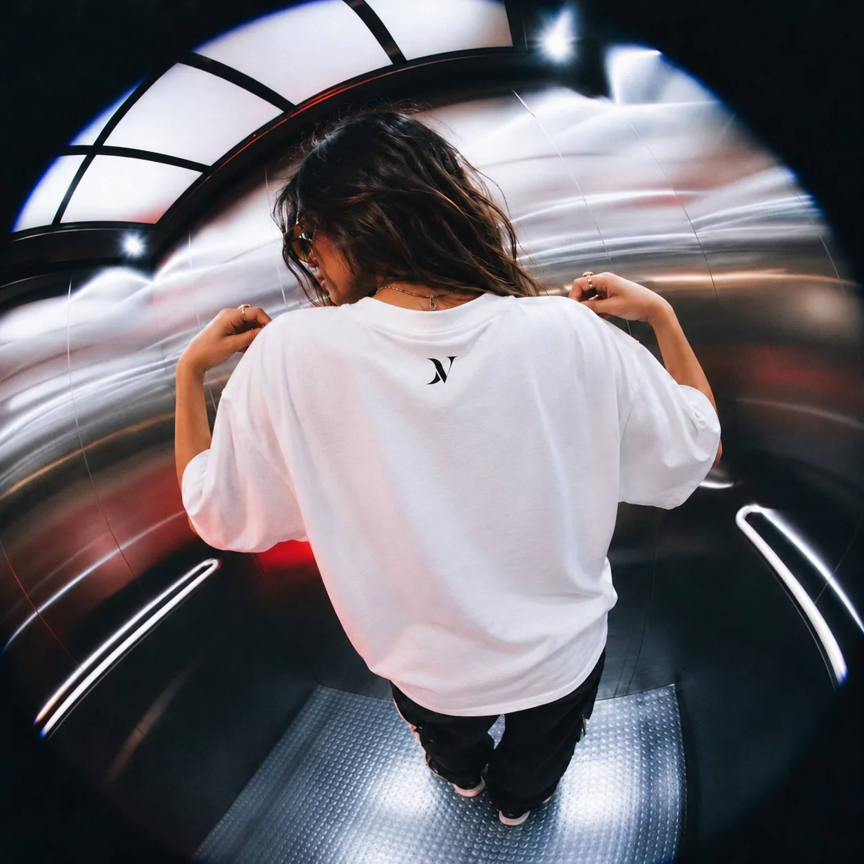 Le dos d’un T-shirt DRAVELL blanc au petit logo noir, porté dans un ascenseur à miroirs.