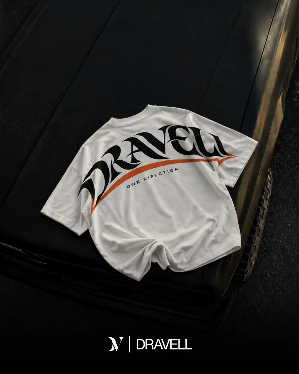 Un T-shirt blanc au grand motif DRAVELL noir et orange posé sur le capot d’une voiture noire.