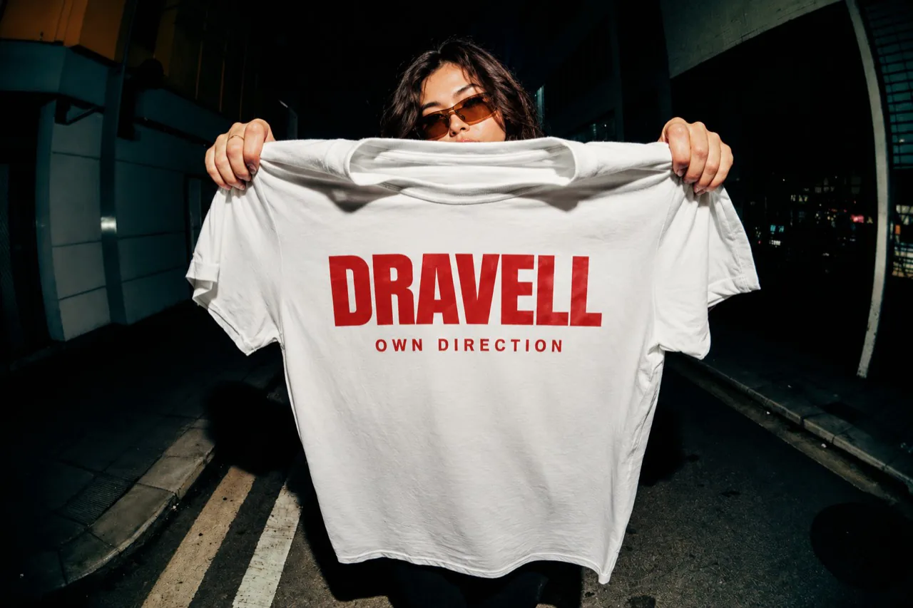 Une femme présentant un T-shirt blanc au lettrage DRAVELL rouge dans une rue.