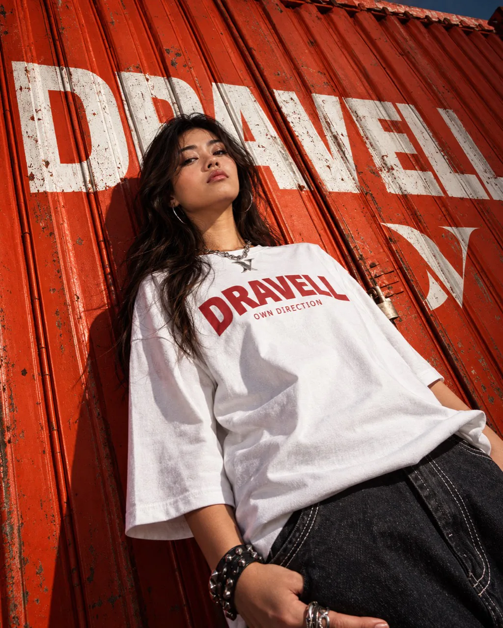 A woman wearing a white DRAVELL T-shirt in front of an orange shipping container with a large DRAVELL wordmark.