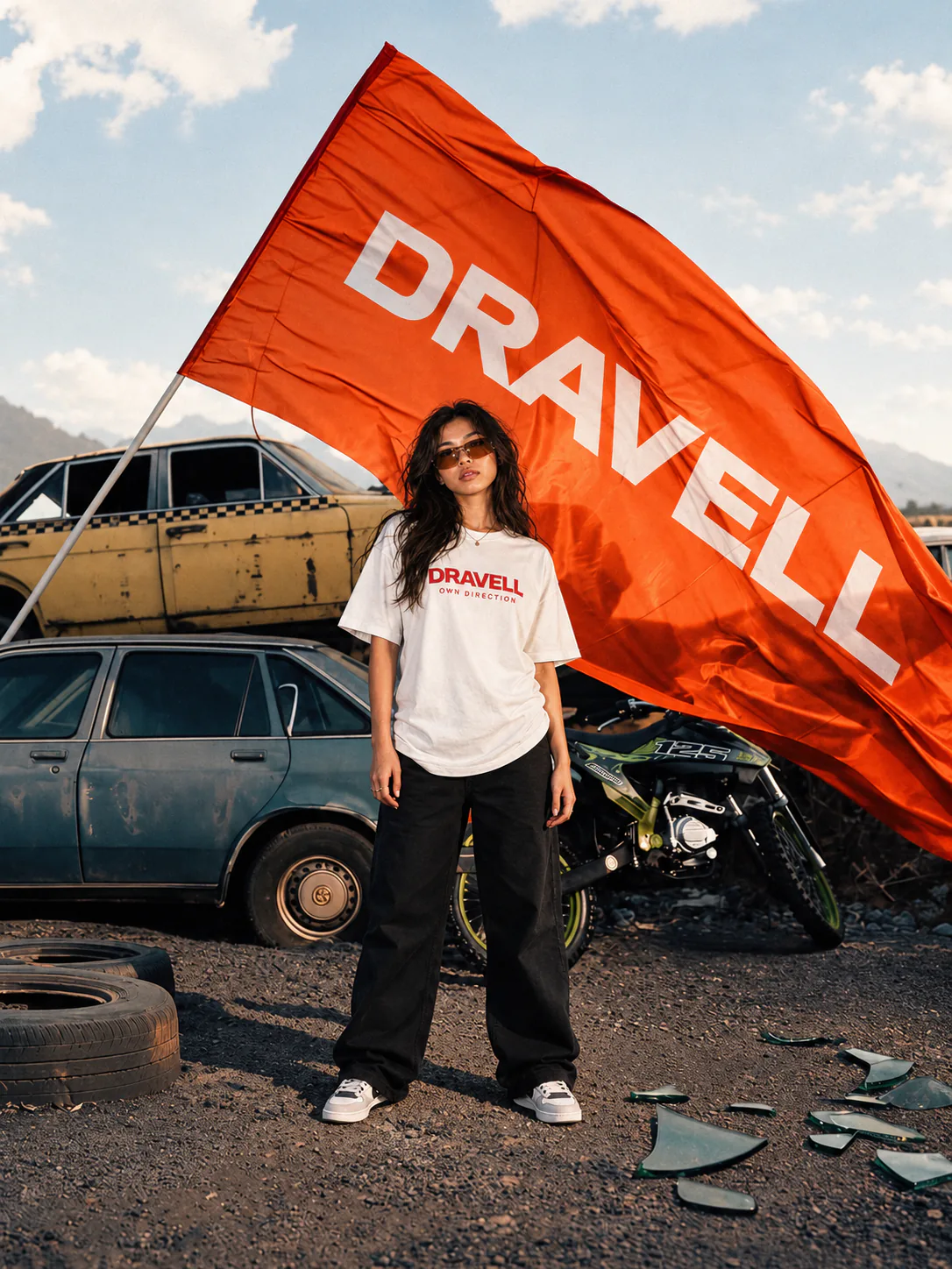A model in a white DRAVELL T-shirt beside stacked cars and a motorcycle, beneath a large orange DRAVELL flag.