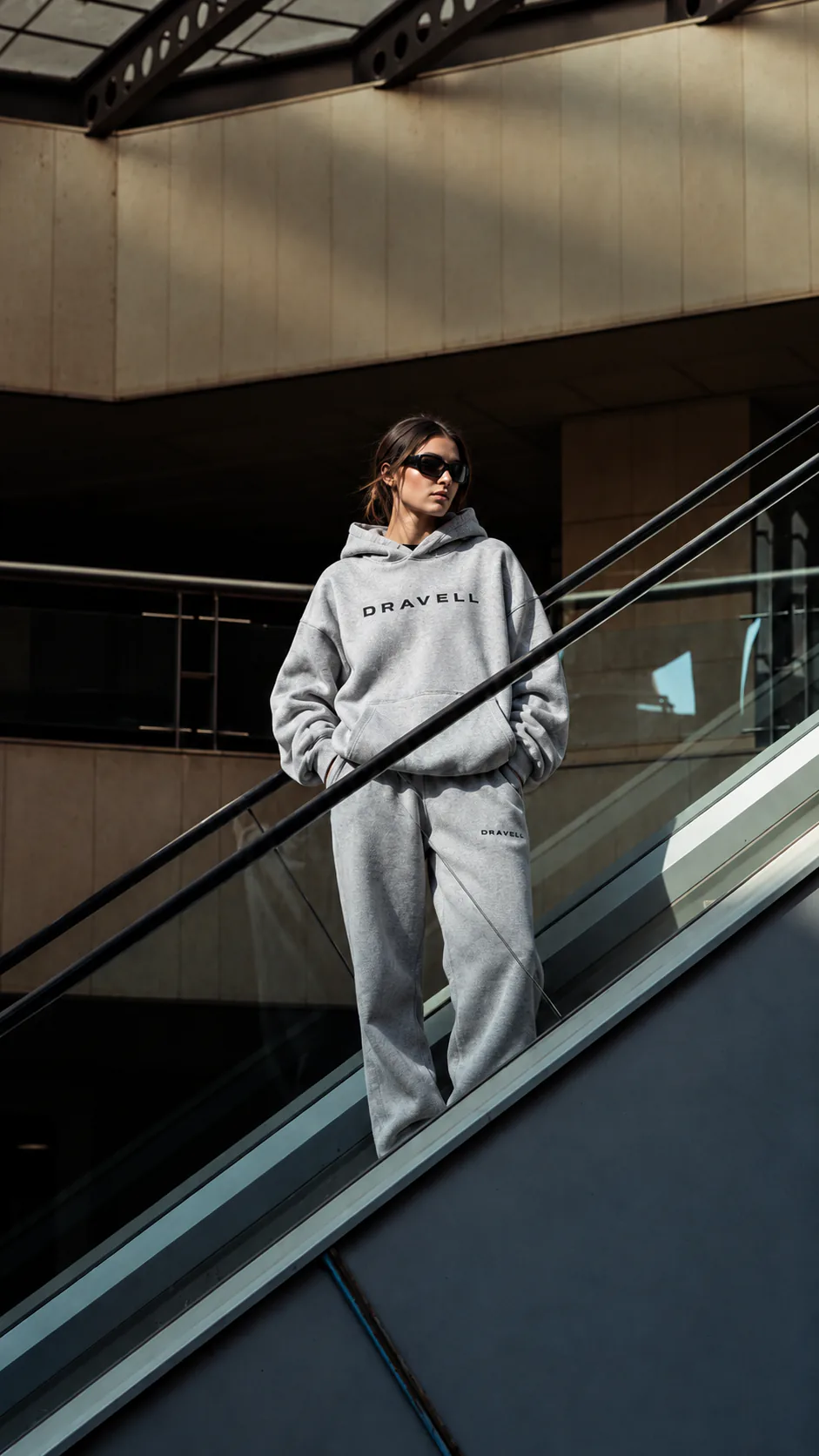A model wearing a grey dravell hoodie and matching trousers on an escalator.